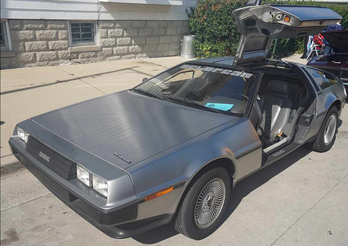 A silver car made of stainless steel with a long, boxy hood and a driver’s side door open. The door opens from a hinge at the top of the car so that it looks like a wing rising above the top of the car.