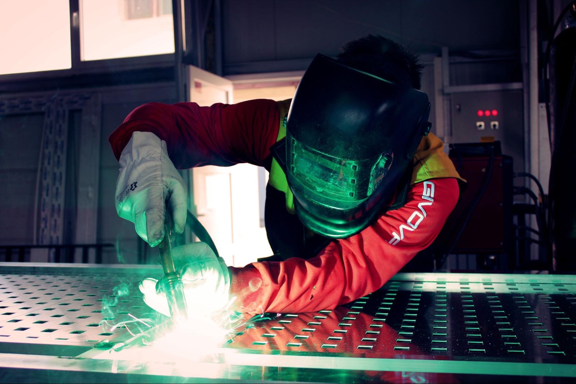 A person wearing protective equipment welds a long aluminum fixture.