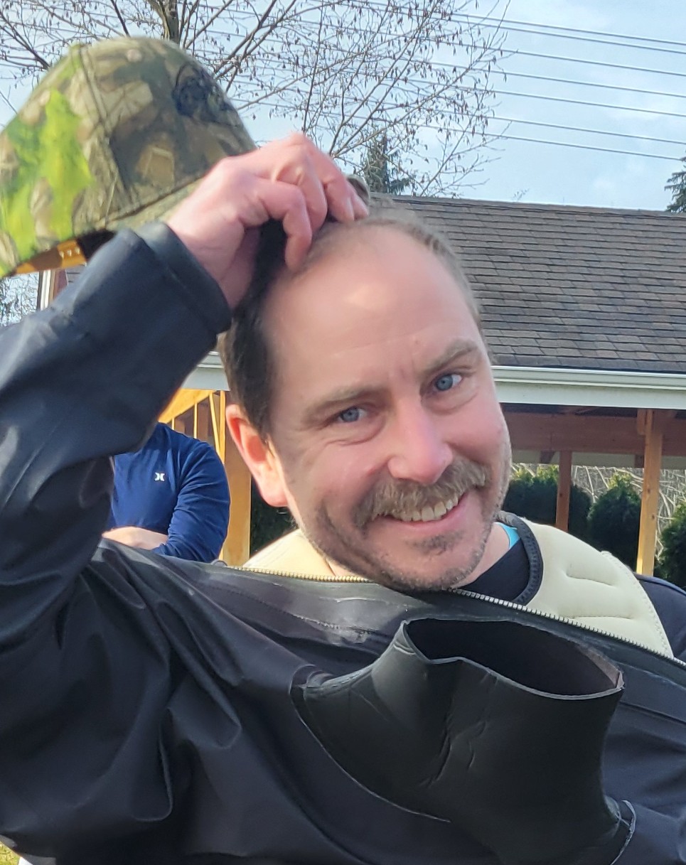 A white man wearing a wetsuit smiles into the camera as he takes a ball cap off of his head.