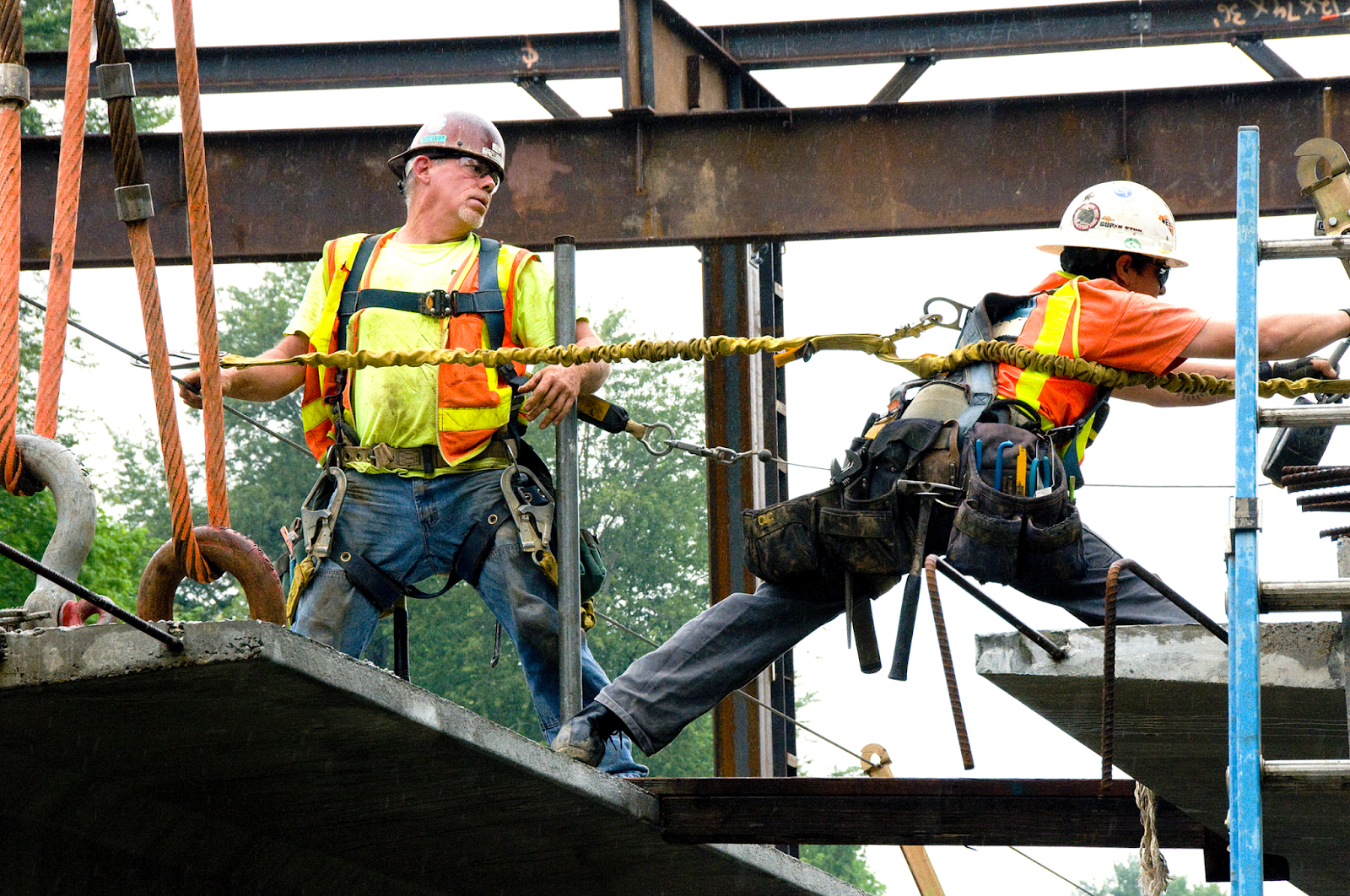 Several individuals work on a building. One person reaches across a platform to secure his lanyard to a second position.