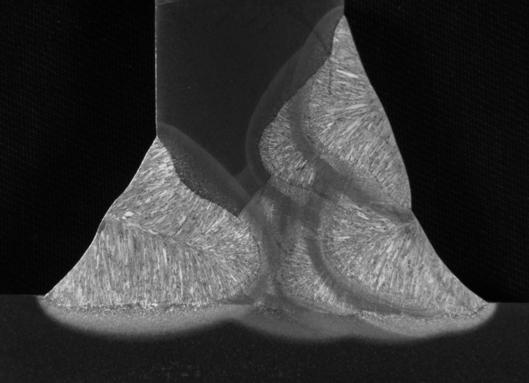 Macroetch of a T-joint is shown where a weld joint has been cut perpendicular to the direction of welding to expose a cross section of the weld beads. This surface is then treated with an acid solution to highlight the differences in the microstructure of the metal. This image has been enhanced with special lighting contrasts to show the heat affected zones and the individual grains in the weld metal.