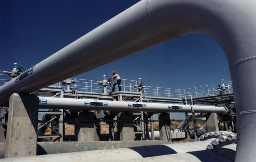 Oil Piping showing a large pipe that starts in the lower right of the image, goes up the right of the image, and then curves to reach across the top of the picture as if framing the worker behind the pipe. In the frame, a worker turns a valve that controls the pipeline.