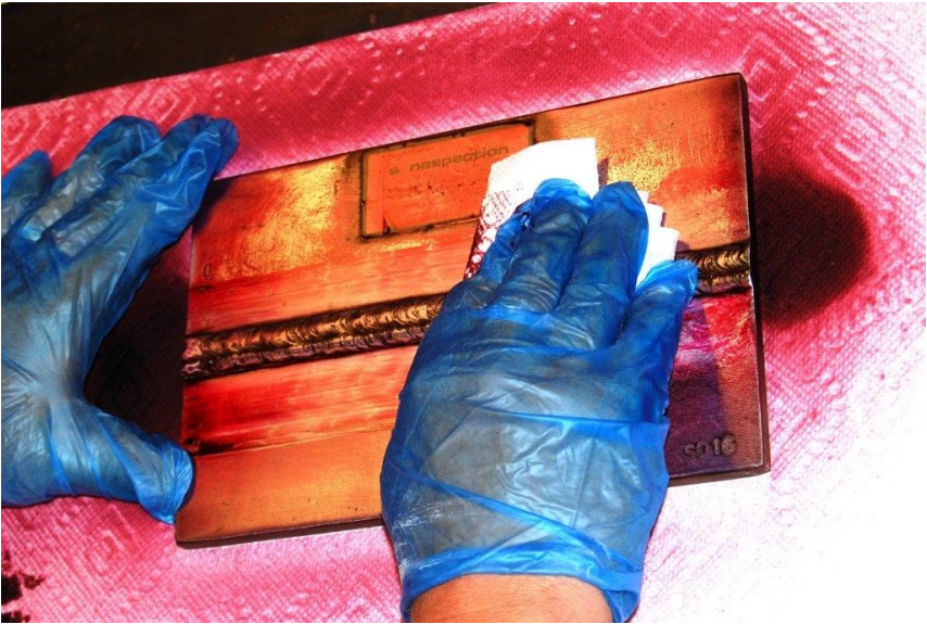 A close-up of two hands, wearing latex-style gloves using a paper towel to clean the surface of a weldment covered in red liquid penetrant.