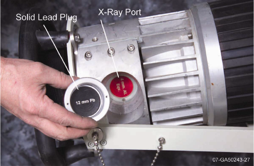 Radiographic X-Ray generator and the solid lead plug removed. A large cylindrical device with a hole in the top part of the cylinder that is labeled X-Ray port. A hand holds the solid lead plug, which is also labeled. The plug caps the X-Ray port when the rodiographic x-ray generator is not in use.