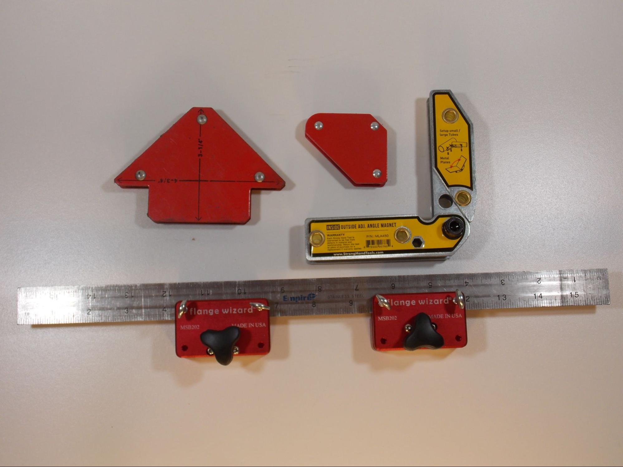 Various types of magnets for use in welding. One is shaped like an isosceles triangle, one is shaped like a right angle, one has two bars connected with a screw so that it can be opened and closed to the appropriate angle. The final magnet is a ruler with two red rectangular magnets about one quarter of the way from either end of the ruler.