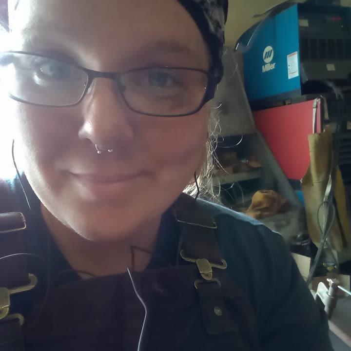 A smiling woman wearing welding gear, glasses, and a nose ring in a shop environment.