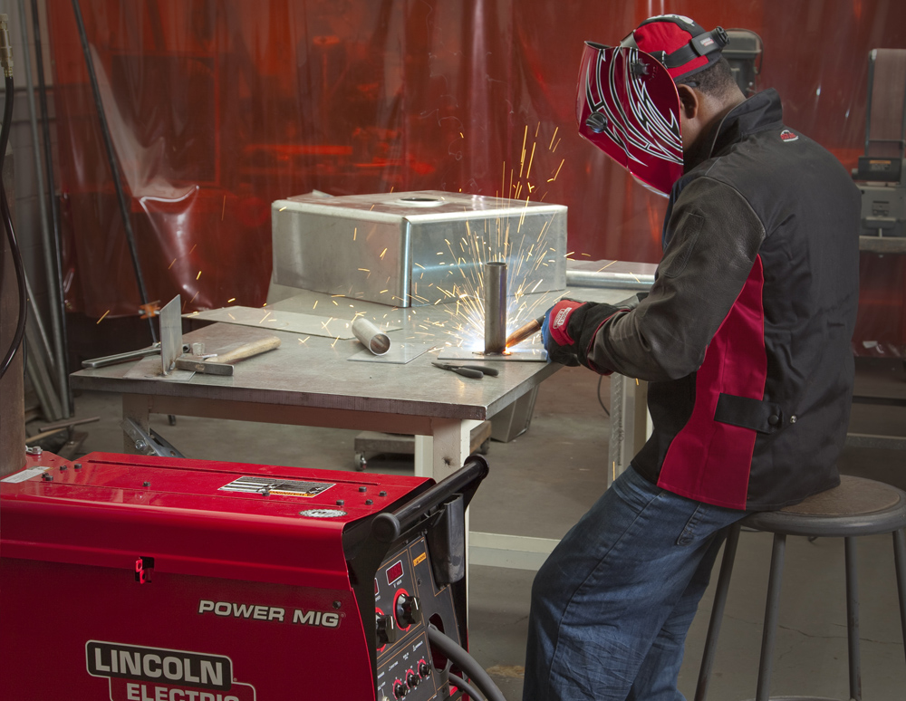 A welder in a black and red welding jacket and hood sits at a work bench welding a tube to a plate