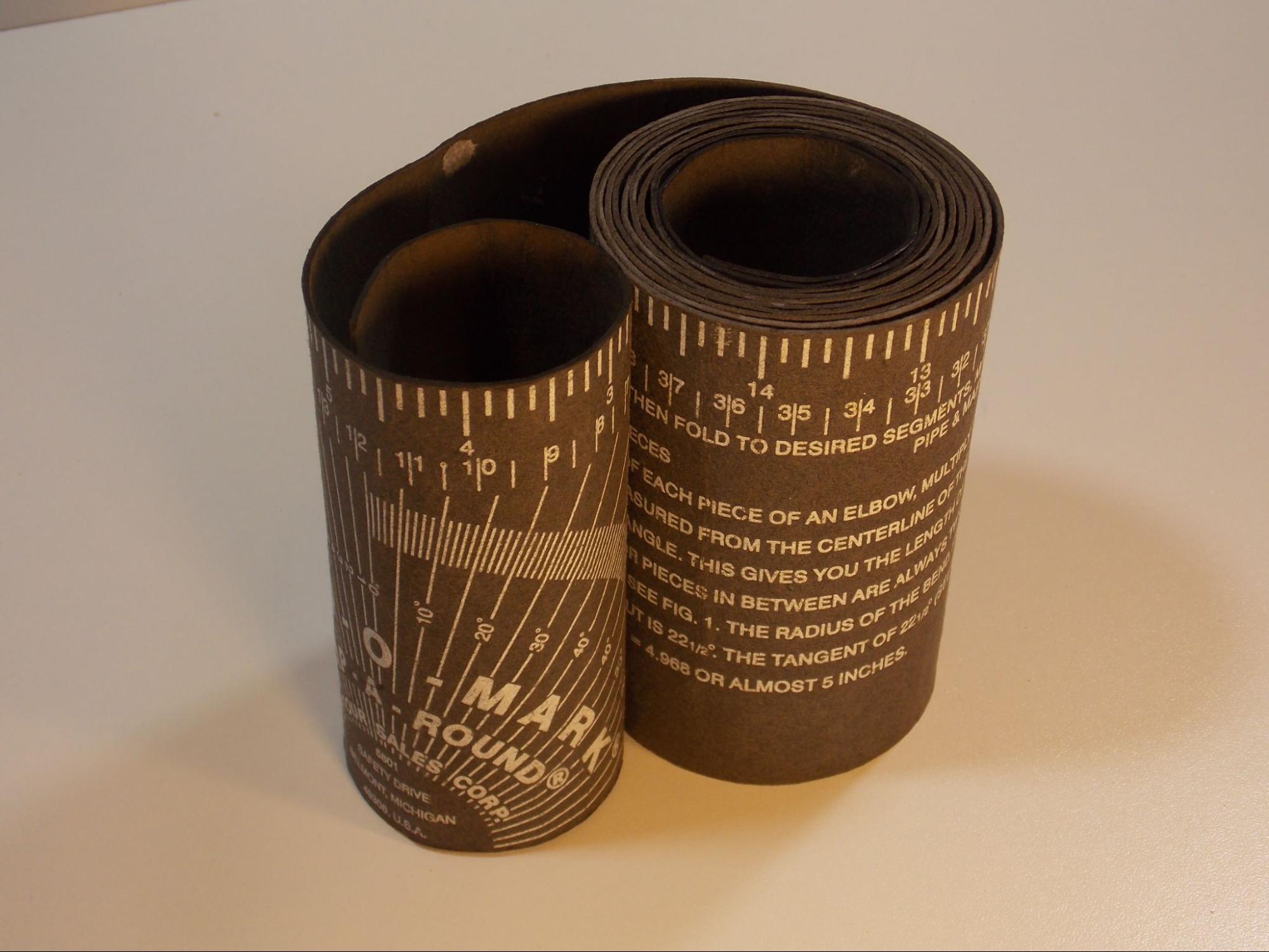 A picture of a rolled up pipe wrap, which is a long strip of pliable cardboard with the Imperial measurement scale printed on the top edge and a 180 degree scale presented on the flat face of the tool.