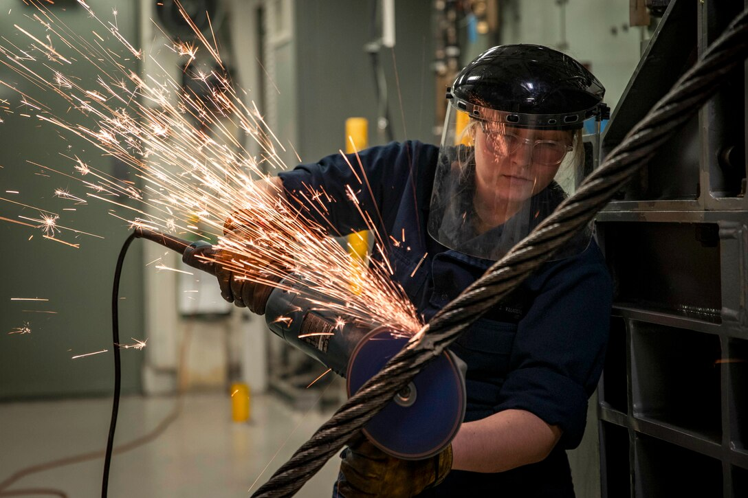 A worker uses a large handheld angle grinder to grind welds on a wire rope.