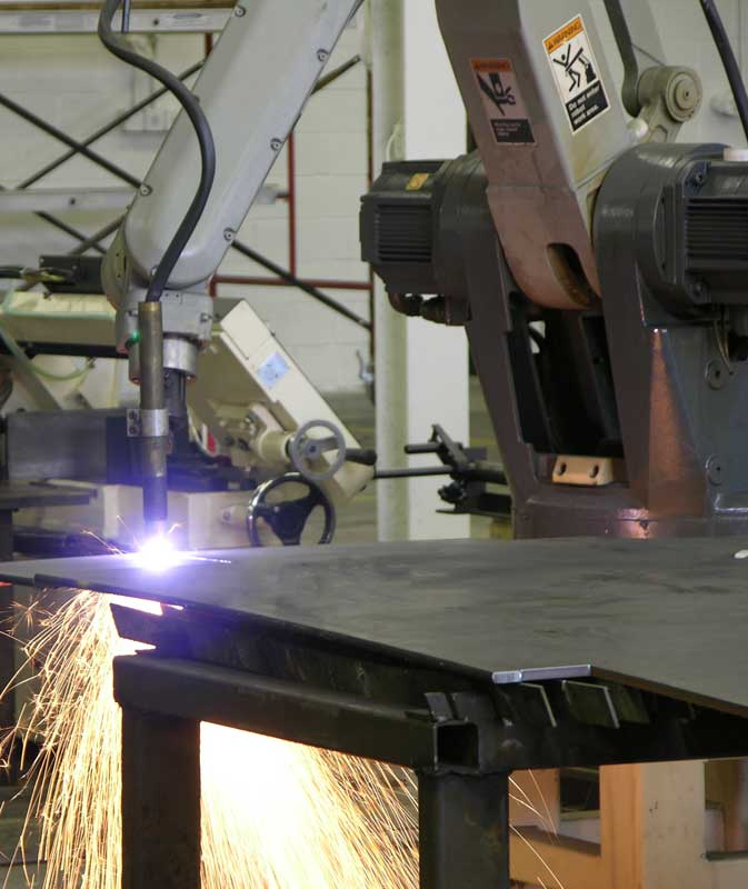 A plasma torch, mounted on a robotic arm, makes a cut on a large flat piece of metal that is laid out on a table.