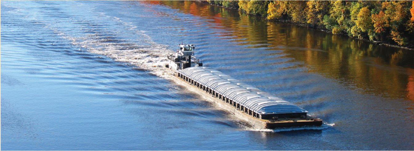 a barge traveling close to the shoreline. The barge is being pulled by a tugboat and has a cargo lifted on pallets.