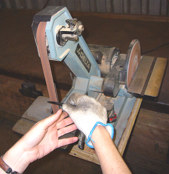 A small tabletop belt sander. The machine is light blue, with the brand name Delta printed on the front. A person wearing a glove holds a piece of metal against an abrasive belt that rapidly rotates as it moves around two drums.