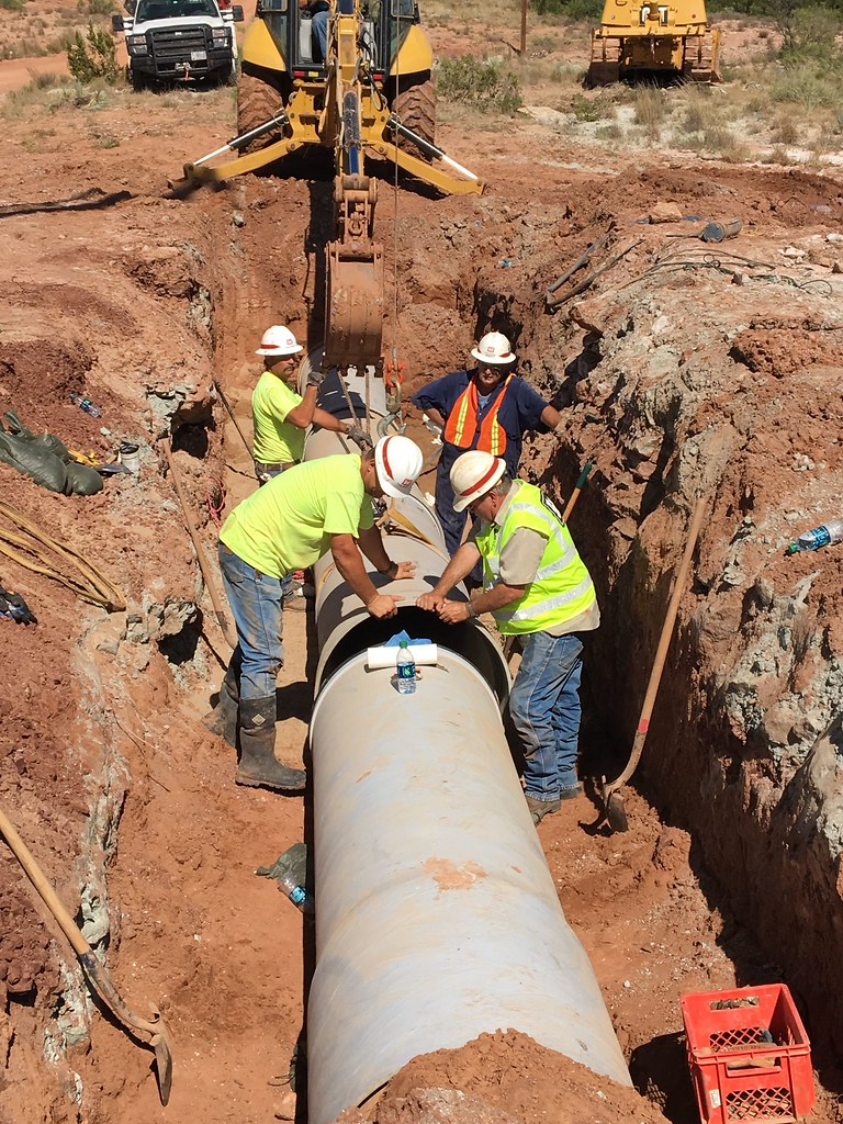 Several workers are in a trench working to align large pipe sections for welding.