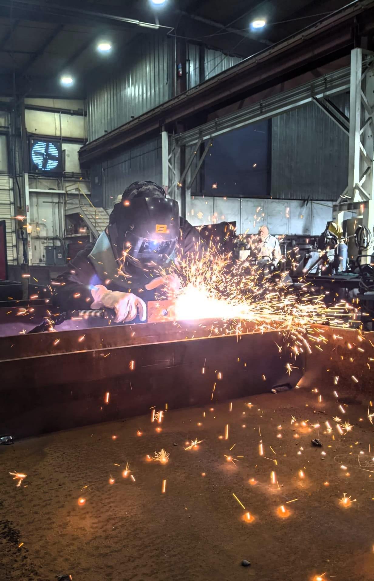 Jessica wearing a welding hood, jacket, and leather gloves uses a plasma cutter to trim a part on a box shaped weldment.