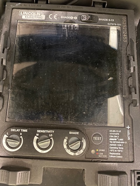 The control panel on the inside of the welding hood. There is a large viewing window, controls for delay time, sensitivity, shade, and a battery test