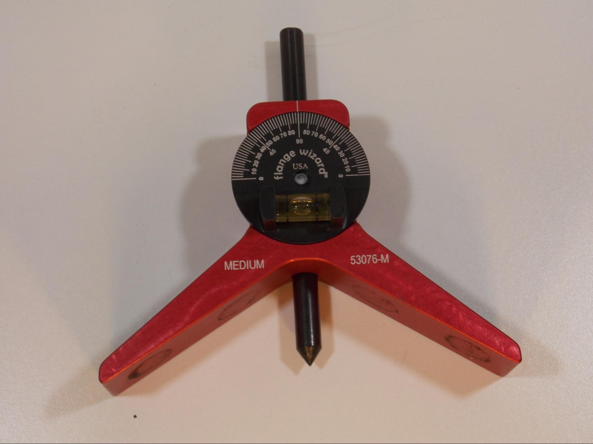 A pipe centering head is pictured. It is a red and black tool with two arms forming a wide angle so that the tool may be placed against a tube or pipe. A black metal spike called a center punch runs through the center of the tool, and a bubble level is affixed to the face of the tool above the two arms. The level has a 180 degree scale.