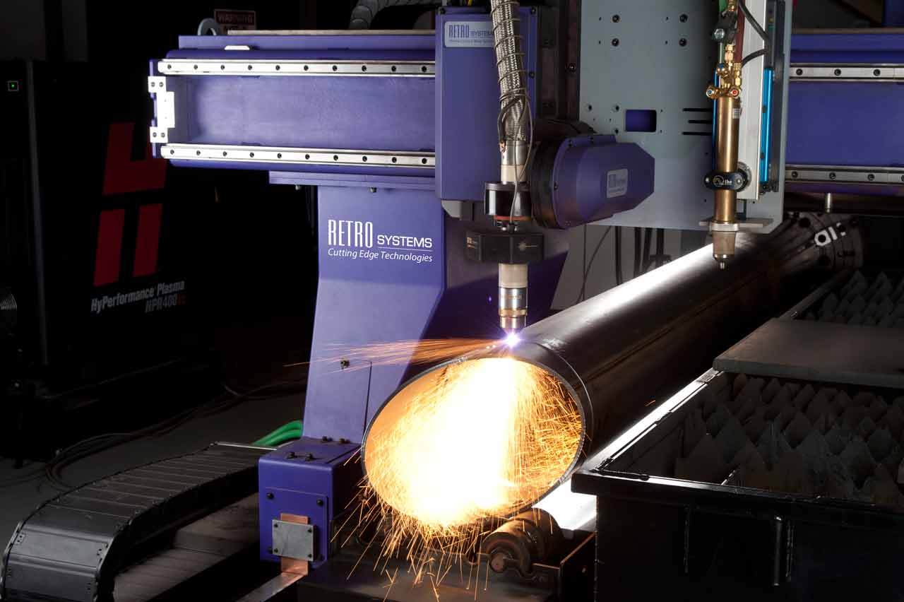 A metal pipe set up on a computer numeric control plasma table. The table has RETRO Systems: Cutting Edge Technologies printed on it. The machine is performing a plasma cut on the pipe.