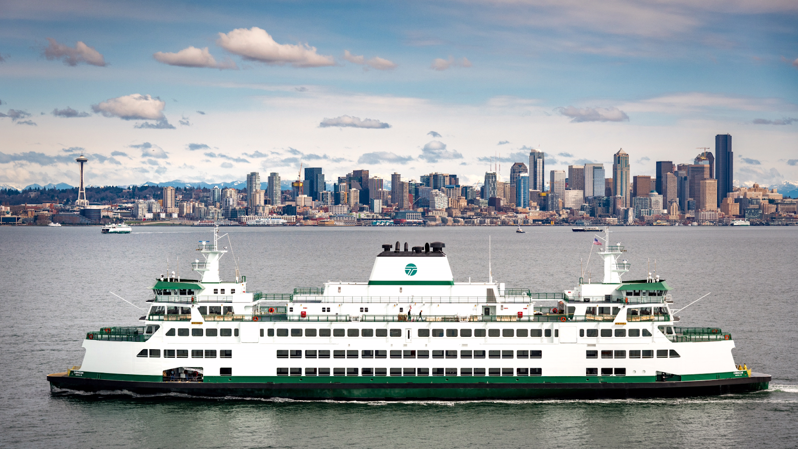 In the foreground is the Washington State Ferry, Chimacum. The ferry is mostly white, with green detailing at the waterline and on railings. In the background is the city of Seattle showing tall buildings along the skyline.