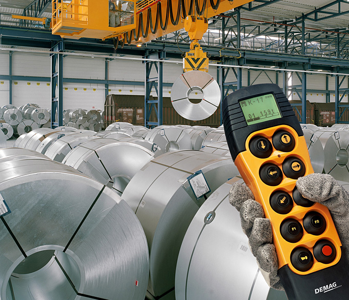 An overhead bridge crane lifts huge, heavy coils of steel in a warehouse. A worker holds a remote control unit for the crane.