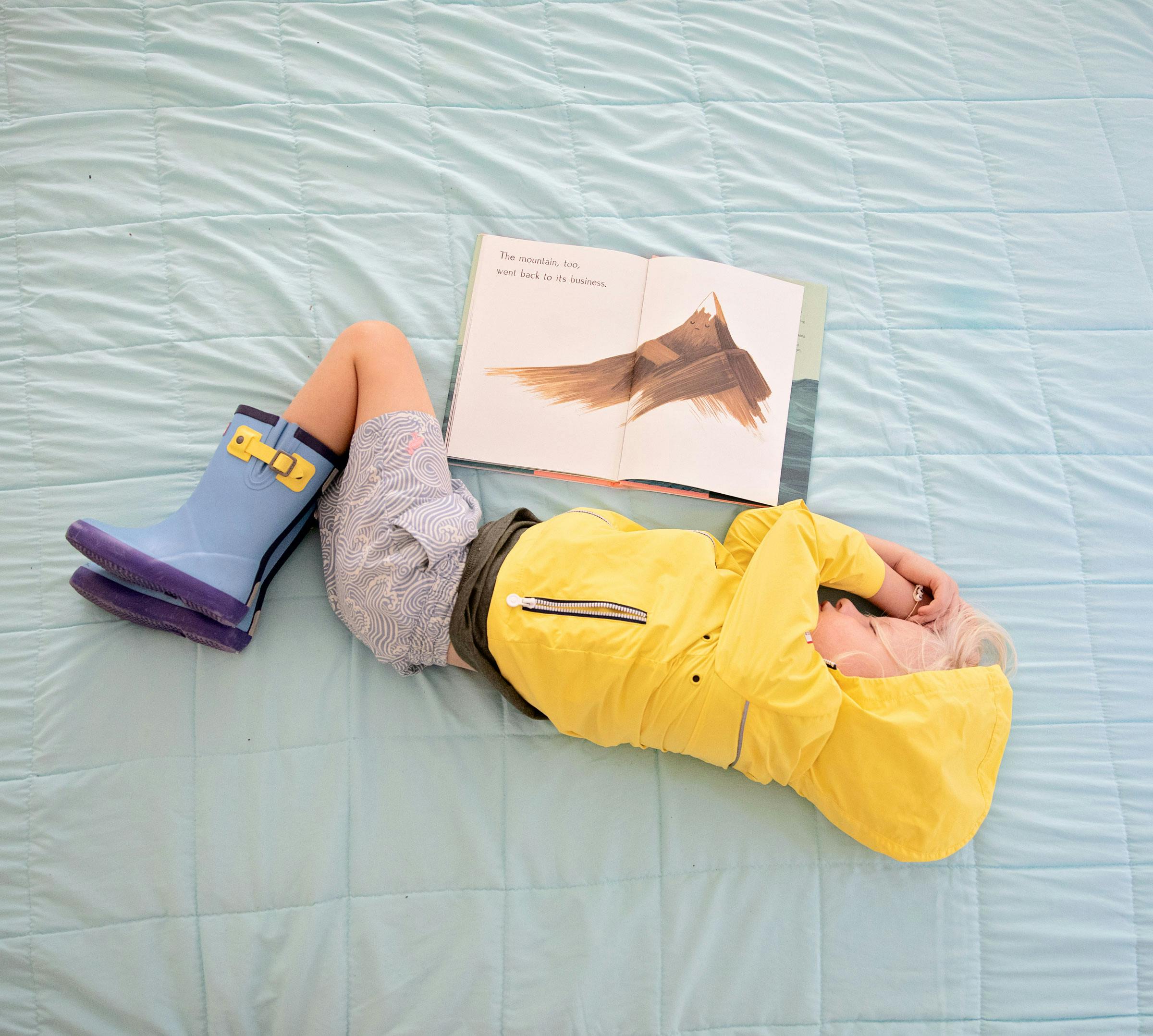 Child napping in raincoat and boots next to open book.