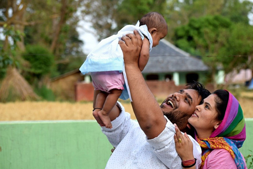 Man holds a baby up in the air, smiling. Woman stands next to the man, also smiling at the baby.