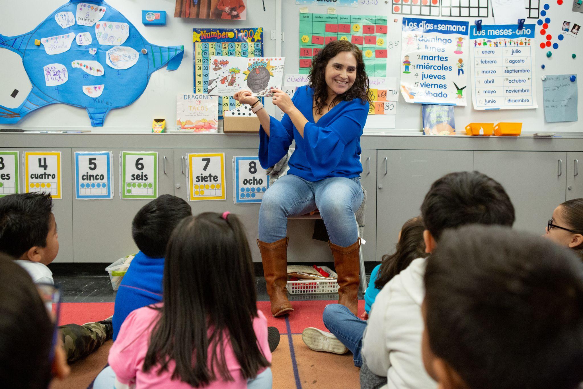 A female teacher sits on a chair, holding a picture book for children seated on the floor.