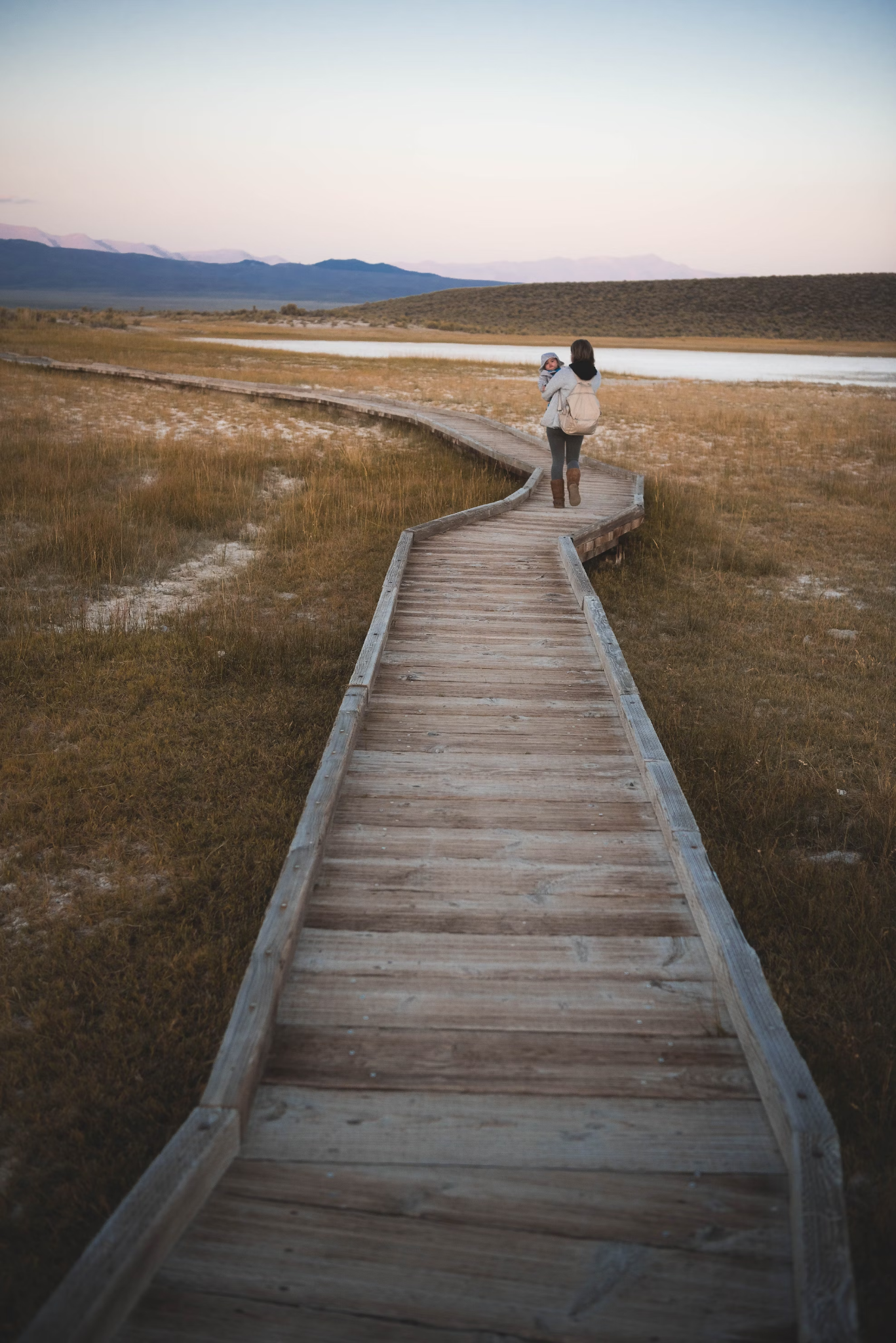 Parent holding a young child and standing on a very long wooden walkway outside, looking off into the distance, indicating possible challenges or barriers.