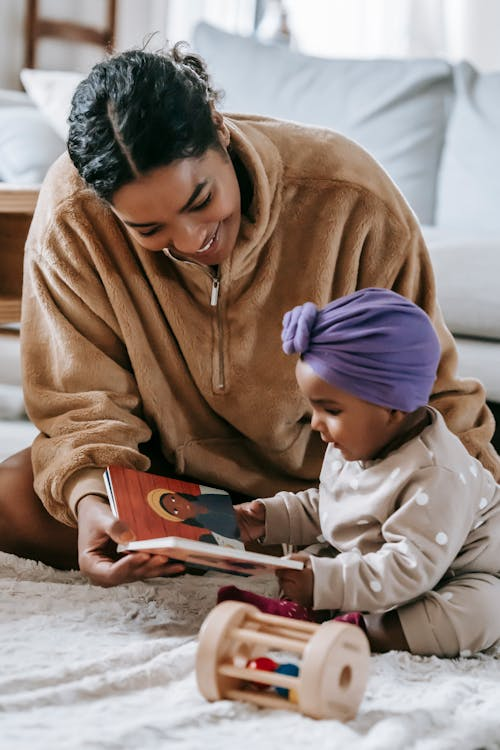 Mom sitting on the floor reading a picture book with her daughter. The book is meant to be a racially-appropriate and developmentally appropriate choice for the child as the mom reads and talks about the pictures.