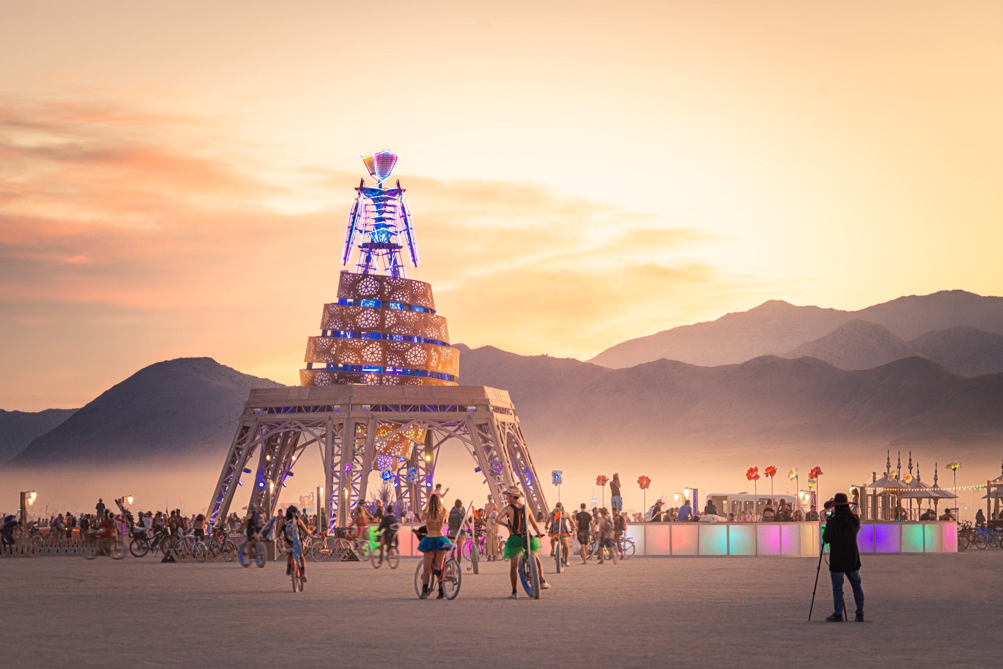 Photograph depicting a multistory tall art structure that resembles a skeleton standing atop a platform with blue lights highlighting artistic designs. The structure is placed on a flat sandy expanse with mountains in the distance. Many people are standing, riding bicycles, or photographing the sculpture.