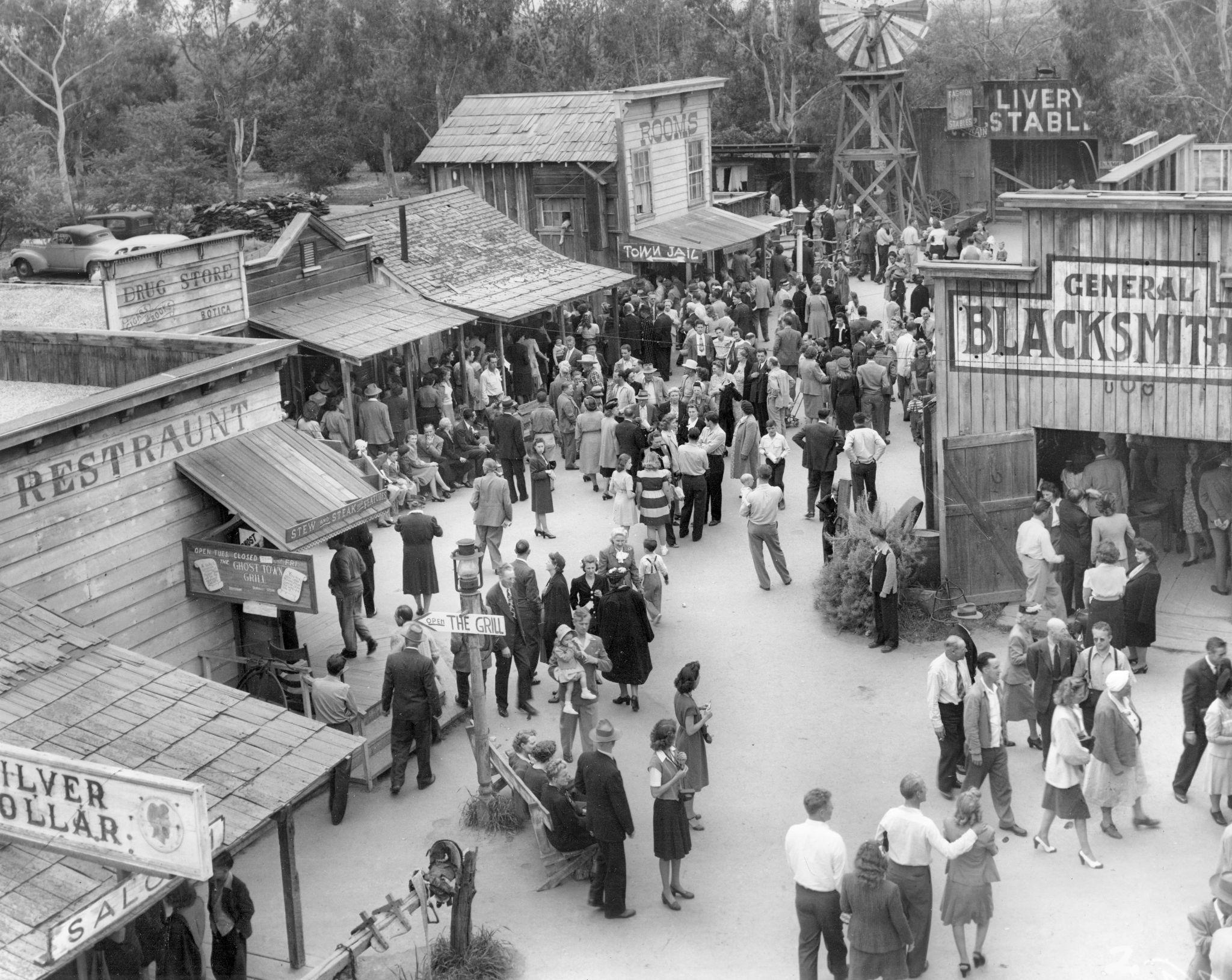 Monochrome photograph showing a crowd of people milling amongst wooden structures reminiscent of a historic western town.