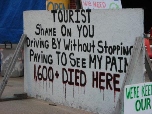 Hand-written sign held up by wood braces reading "tourist, shame on you driving by without stopping paying to see my pain 1600+ people died here"