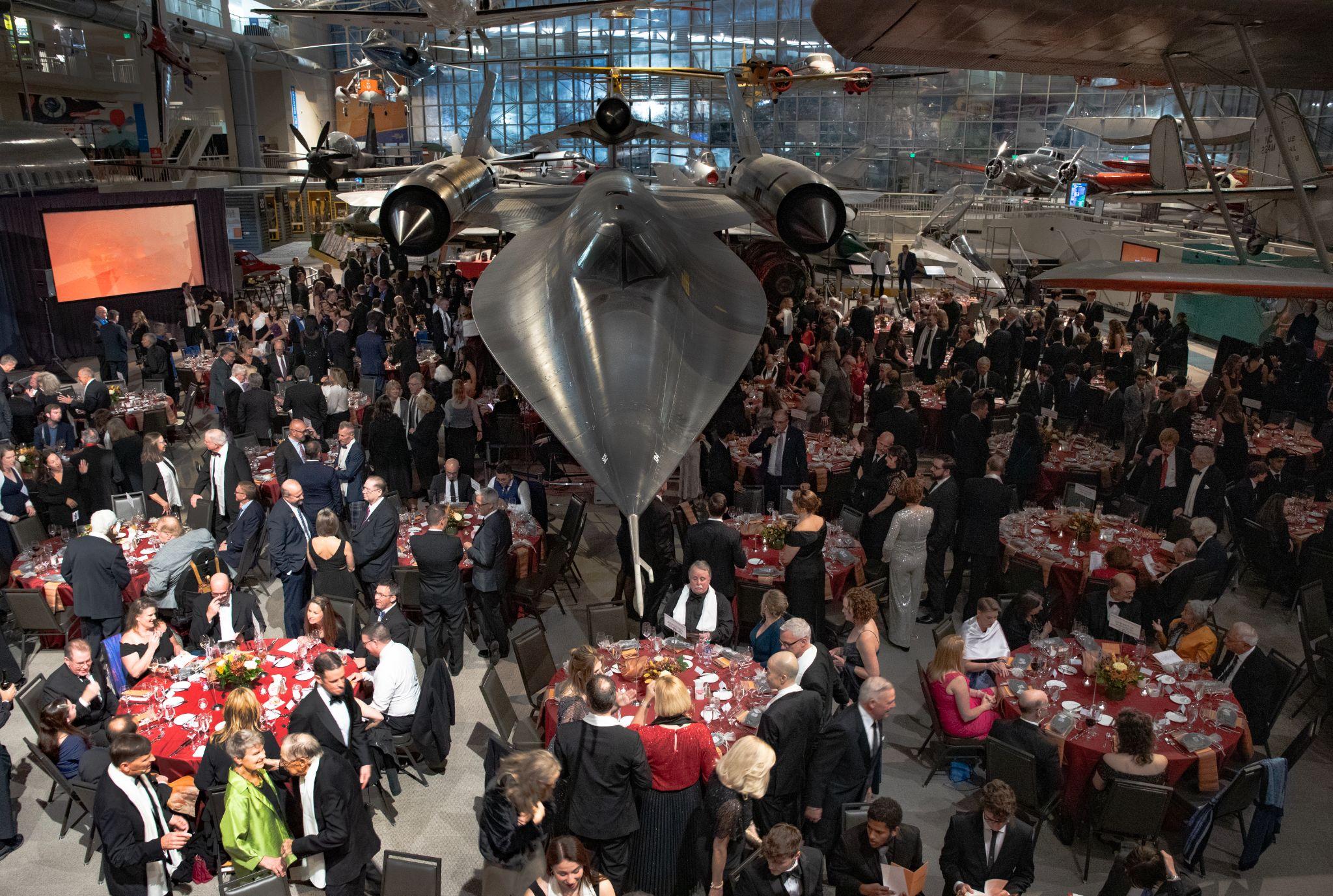 Many people, men and women, dressed formally and sitting at round dining tables. The tables are situated underneath the SR 71 Blackbird.