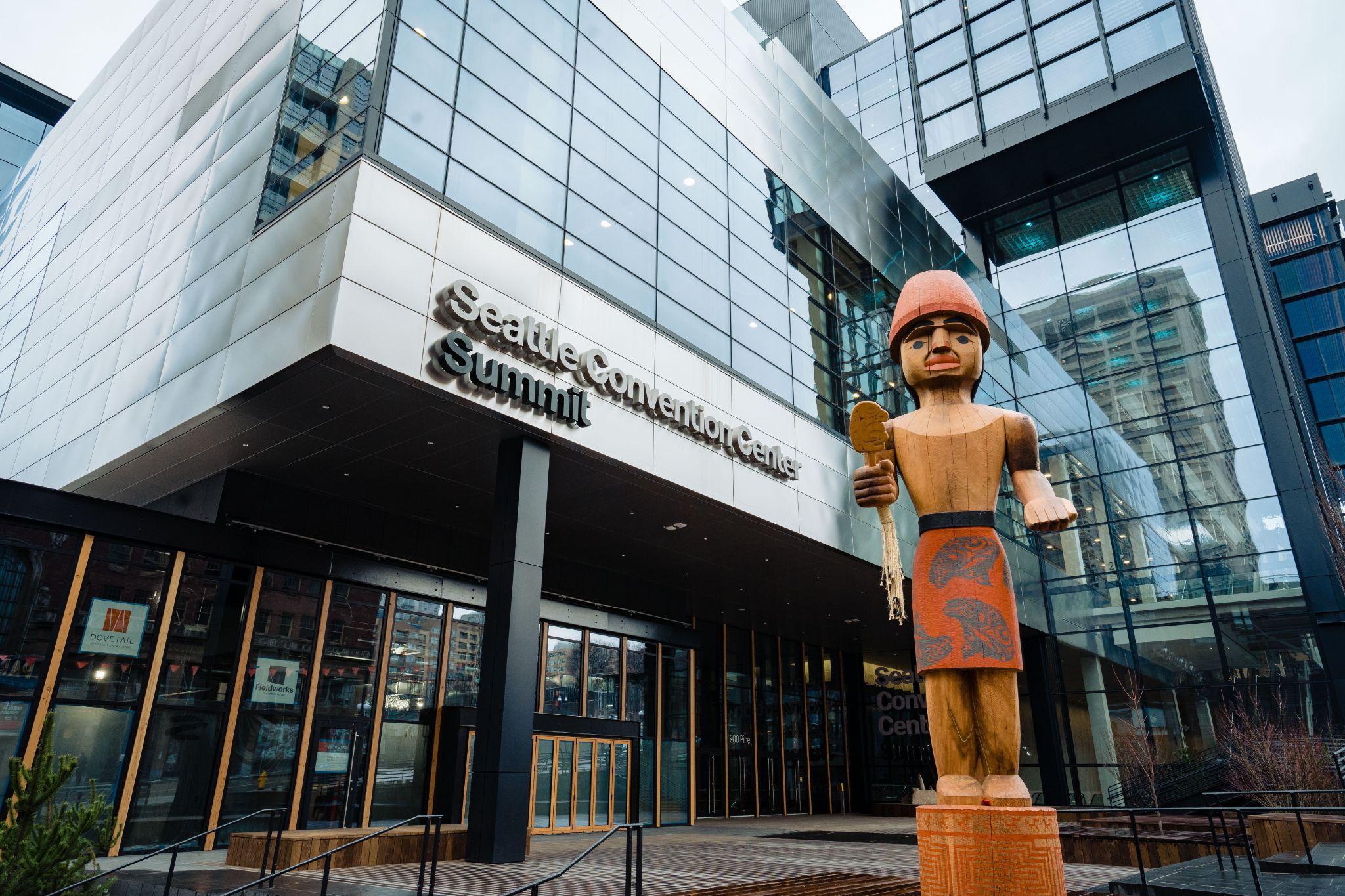 The main entrance to the Seattle Convention Center Summit building, a large modern glass structure. A carved wooden statue of indigenous design stands in the forefront.