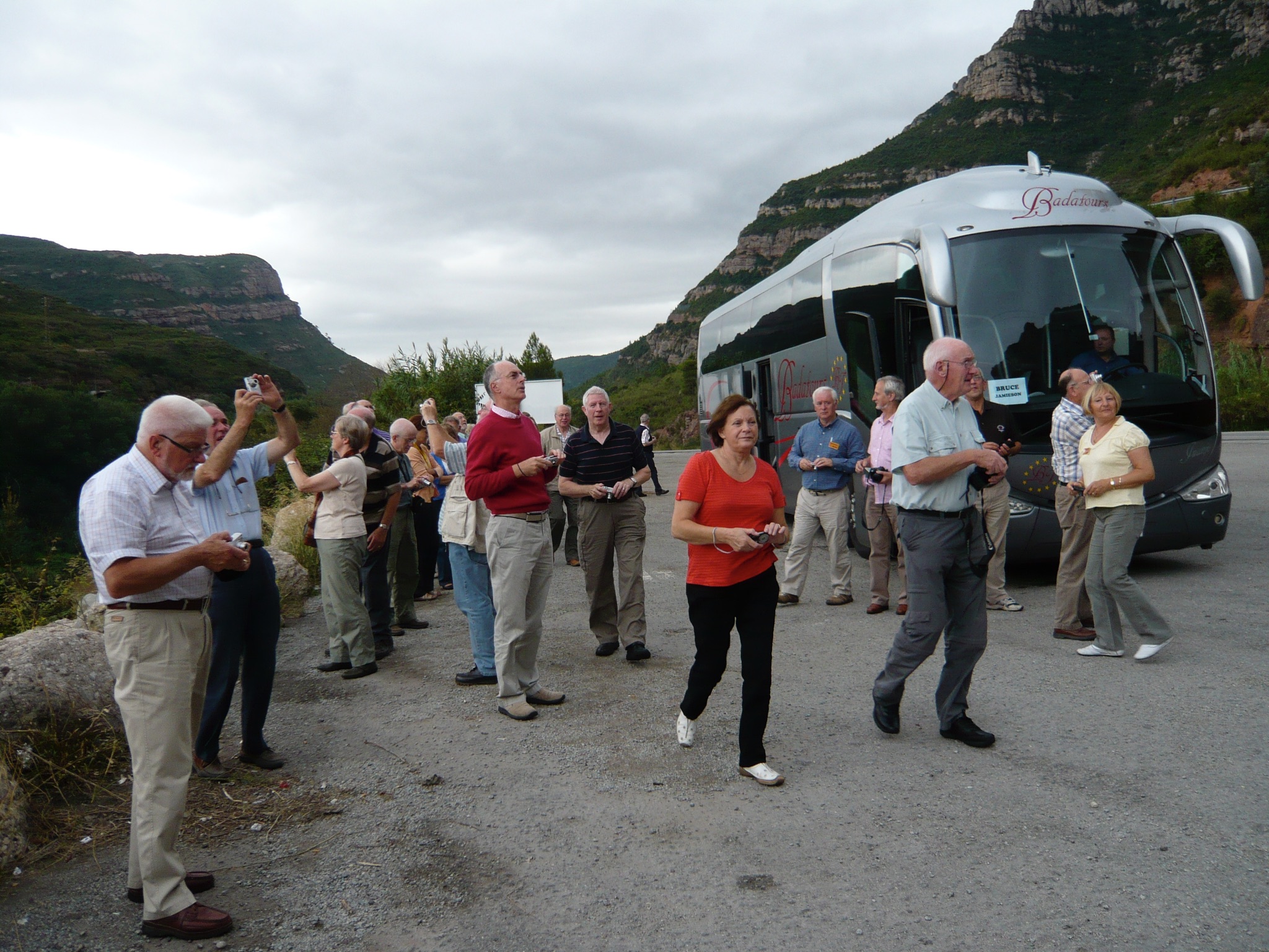 A photograph depicting people gathered on the side of a road next to a motorcoach bus, many with cameras. The landscape is of tall green mountains with rocky outcrops.