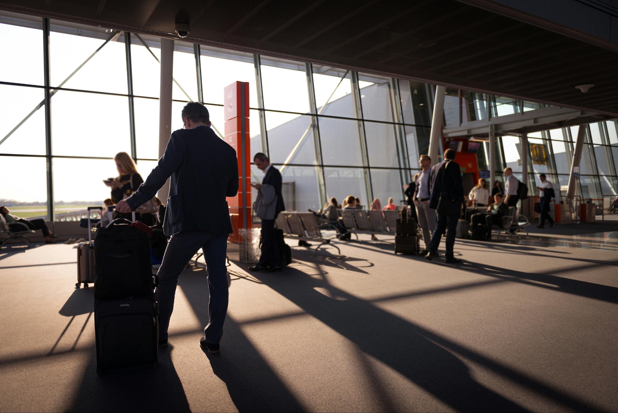 people inside an airport waiting for their flight. The sun is setting, creating shadows. Many people have suits on and have luggage.