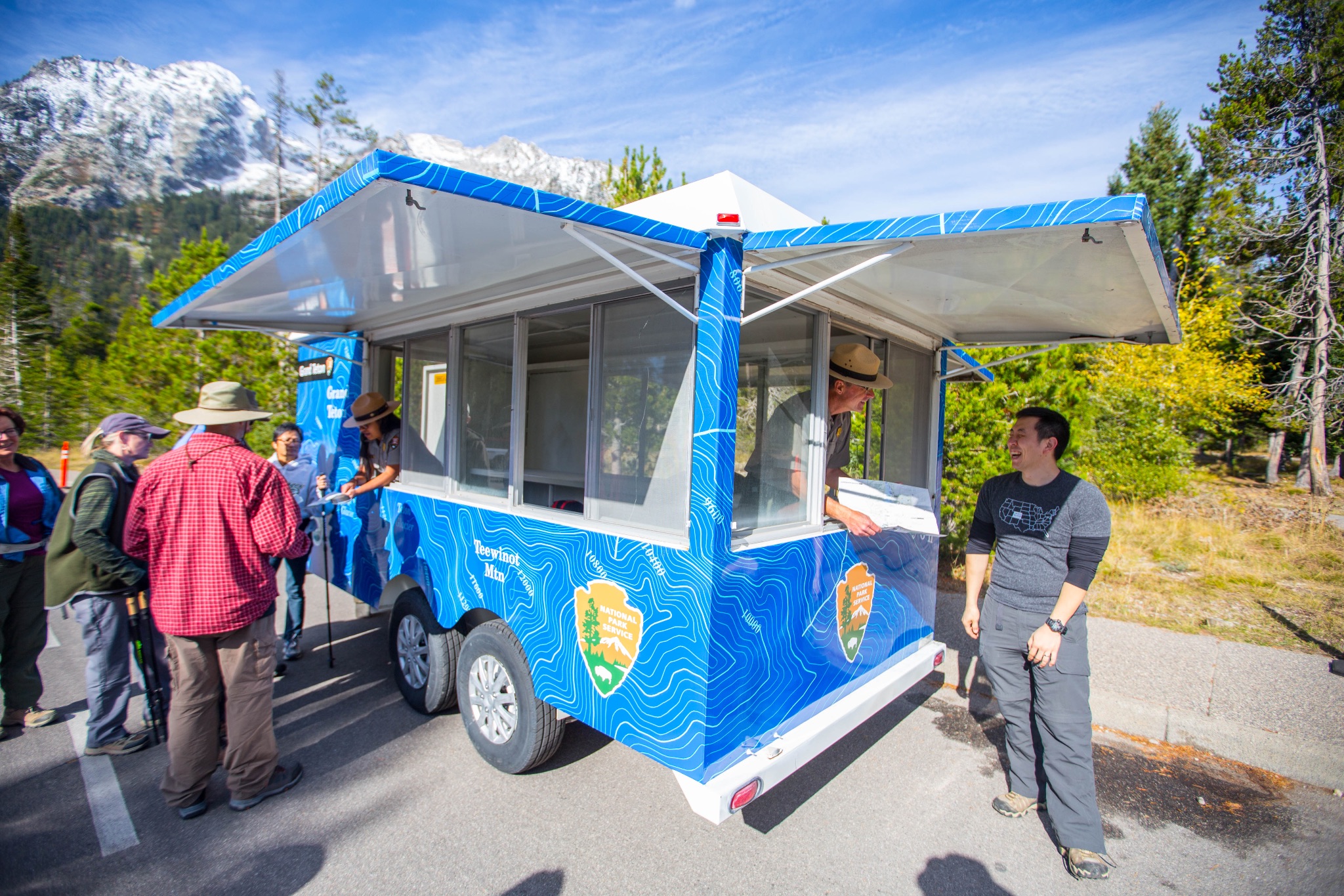 A photograph of a blue, wheeled trailer in a parking lot with its sides opened up to reveal windows and park rangers. Two park rangers are speaking separately with people and showing them maps. Snow capped mountains and forests are in the background.