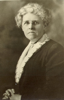 Photograph of Lola Baldwin looking professional, wearing wire-rimmed glasses and a long-sleeved dress, with her hair held back, and sitting with folded hands.