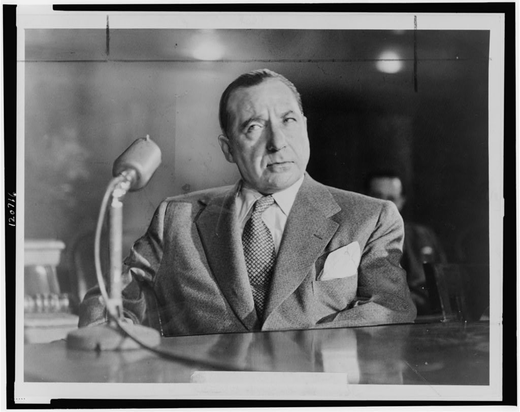 Photograph of the conspicuous Frank Costello, looking dignified in a suit and tie with a prudent expression, seated at a desk, with a sturdy microphone on the table to his right.