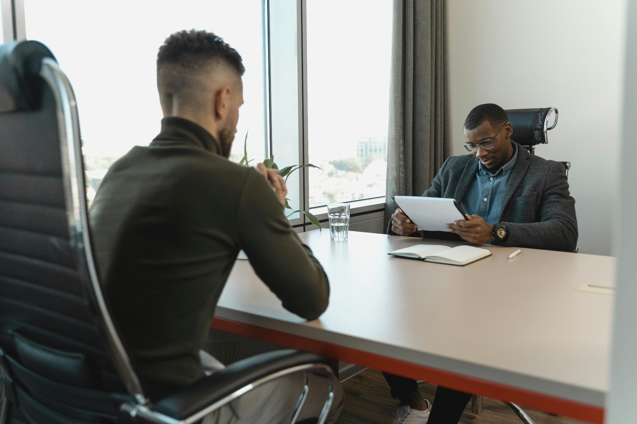 Two men sit at a table opposite one another. One gentleman looks at a tablet he is holding, while the other is sitting forward, elbows on the table, as if anticipating a question.