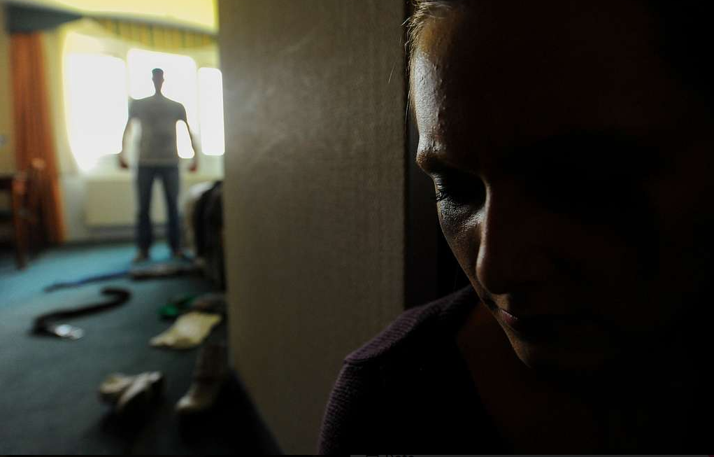 A woman in the right foreground looks down with her eyes closed, the right eye of which is swollen and bruised. In the left background, standing in front of a window, is a standing male subject. Between the two are several items strewn on the blue-carpeted floor.