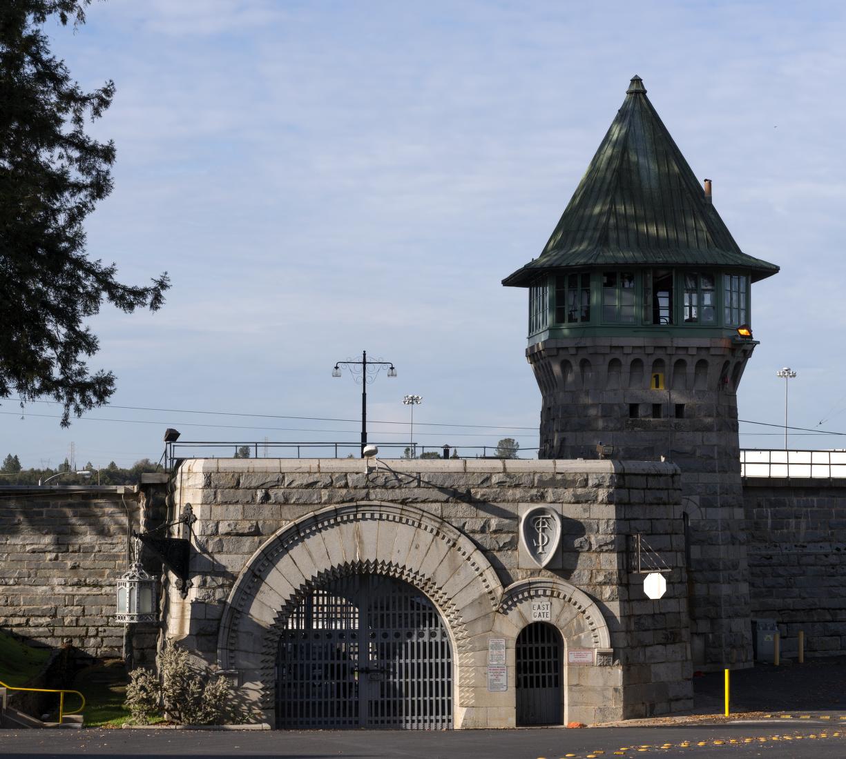 Photograph of the entrance to Folsom State Prison: A stone gateway with a guard tower on top, surrounded by high walls and fences.