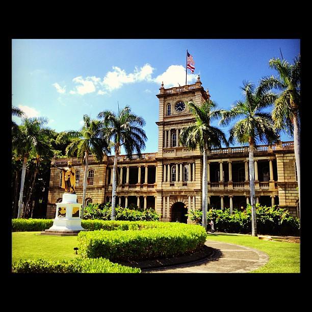 Photograph of the exterior view of the Aliʻiōlani Hale Building, a historic structure located in downtown Honolulu, Hawai’i. The building boasts a grand facade adorned with classical architectural elements, such as columns and intricate detailing. Its notable clock tower rises prominently above the roofline, while the lush greenery and palm trees of the surrounding landscape complement its majestic presence. A magnificent statue of King Kamehameha commands attention at the center of the courtyard.