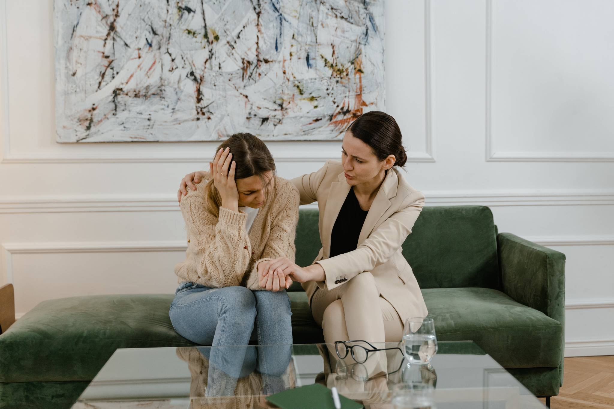 Two women seated on a couch in a white office. The woman on the left is holding her hand to her head and appears distraught. The woman on the right has her right hand on the other woman’s right shoulder, and is holding her left hand so as to comfort her.