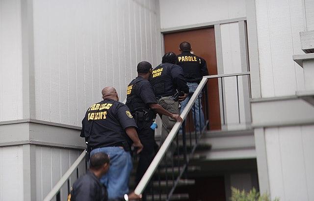 Photograph of five parole officers standing along a stairwell outside the entrance to a residence, preparing to confront an uncooperative parolee.