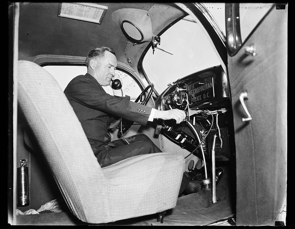 This photograph captures the interior of a Washington D.C. Metropolitan Police automobile in 1936, depicting a man adjusting the dashboard controls while holding the handset of the vehicle's telephone, showcasing the utilization of new law enforcement technologies.