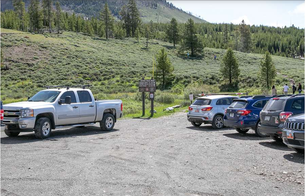 A primitive gravel parking lot with five vehicles, a tree-covered mountain slope and four hikers behind the vehicles. Two regulatory signs are visible at the start of the hiking trail.