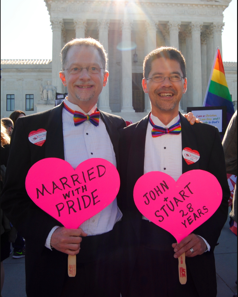 A gay couple demonstrates in favour of same sex marriage about Obergefell v. Hodges in front of the Supreme Court of the United States. The men stand shoulder to shoulder and each hold up pink heart signs. The sign on the left reads “Married with Pride” and the sign on the right reads “John + Stuart 28 years.”