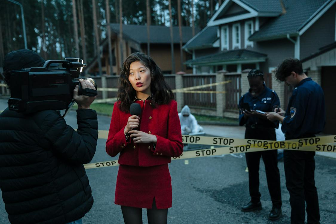 A man operates a news camera on the left foreground, filming a female reporter in a red dress and coat. In the center background is a detective wearing a white proximity suit, in front of two homes and a garage. In the right foreground are two police officers wearing blue uniforms and taking notes.