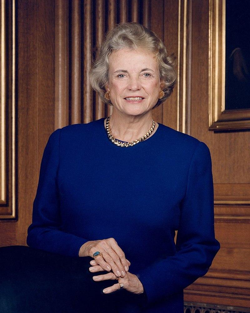 Photograph of Sandra Day O'Connor standing, appearing professional with crossed hands, wearing a conservative royal blue dress and an elegant gold chain necklace.