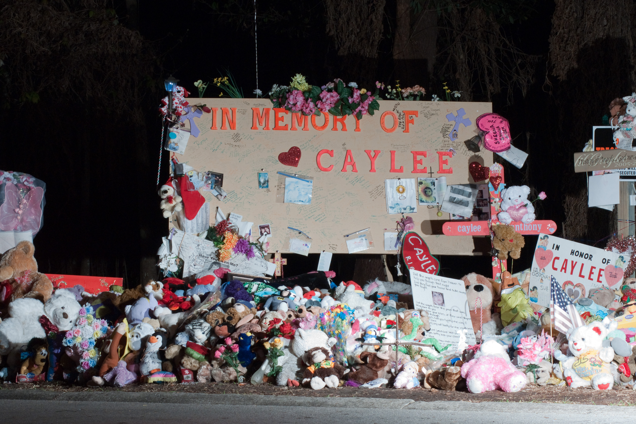 A tan-colored sign with large orange letters reading “In Memory of Caylee” is decorated with drawings, stuffed toys, and flowers. In the foreground are many more stuffed toys, pictures, and a U.S. flag.